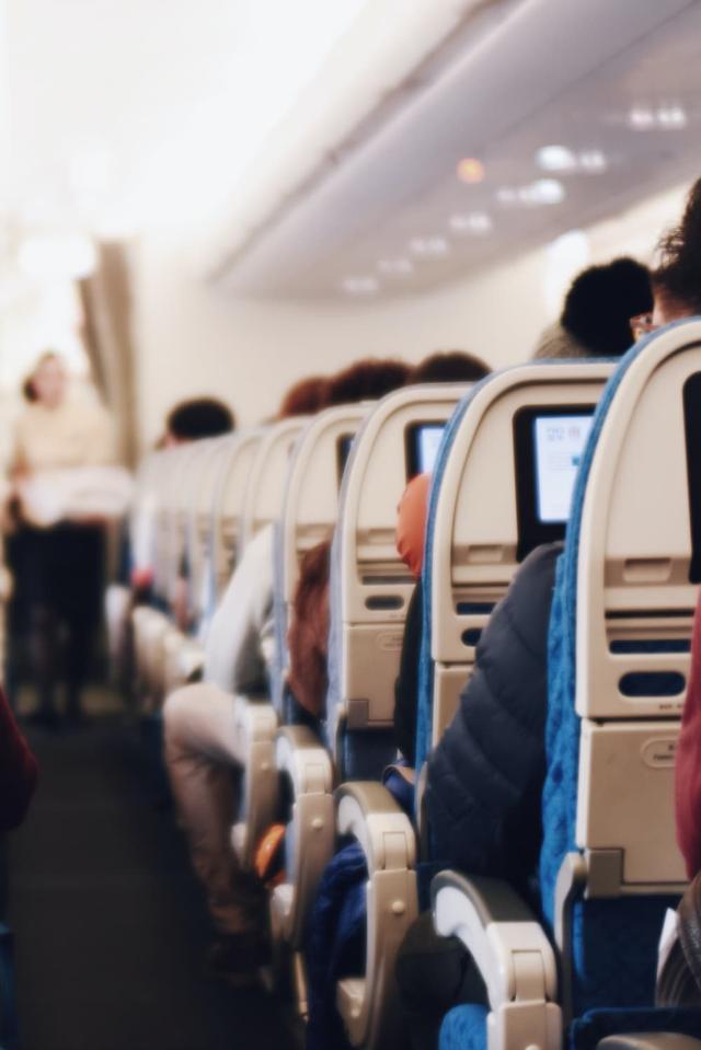 Innenraum eines großen Passagierflugzeugs mit sitzenden Menschen und einer Stewardess in Uniform, die den Gang entlanggeht