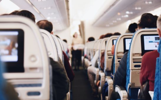 Innenraum eines großen Passagierflugzeugs mit sitzenden Menschen und einer Stewardess in Uniform, die den Gang entlanggeht